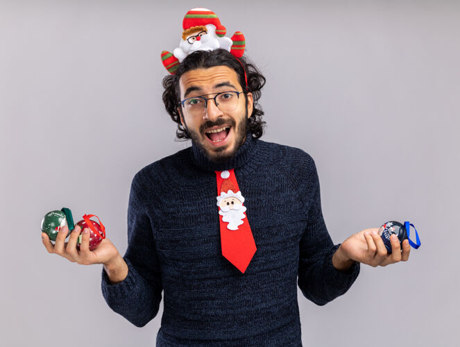 快乐快乐的年轻帅哥戴着圣诞领带 戴着发箍 手里拿着圣诞球 孤零零地站在白墙上穿着帅气圣诞节