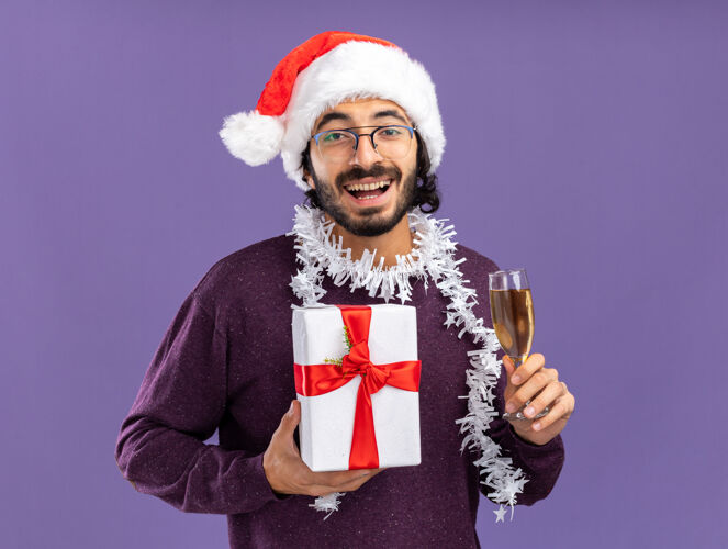 年轻人微笑着的年轻帅哥戴着圣诞帽 脖子上戴着花环 手里拿着一个礼品盒 蓝色背景上隔着一杯香槟礼物脖子小伙子