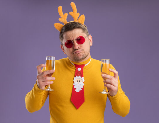 香槟一个穿着黄色高领毛衣 戴着红色眼镜的年轻人 打着有趣的红色领带 戴着鹿角边 手里拿着两杯香槟 站在紫色的背景下 看上去既困惑又不高兴领带角搞笑