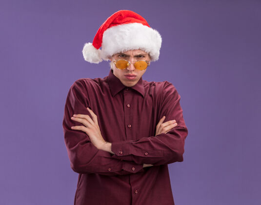 圣诞老人闷闷不乐的年轻金发男子戴着圣诞帽 戴着眼镜 站在一个封闭的姿势 看着被隔离在紫色背景上的相机 还有复制空间眼镜生闷气站着