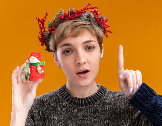 头令人印象深刻的年轻漂亮女孩戴着圣诞花环手持小圣诞雪人雕像指向孤立的橙色墙上小指向花环