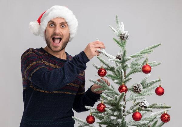 圣诞节令人印象深刻的年轻白人男子戴着圣诞帽站在圣诞树附近触摸它隔离在白色墙上印象深刻男人戴着