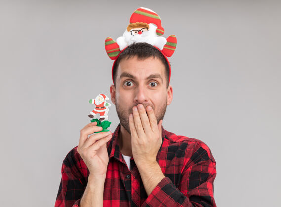 圣诞节关心的白人年轻人戴着圣诞老人的头带 手里拿着雪人的圣诞玩具 手放在嘴上 隔离在白墙上年轻人保管关心
