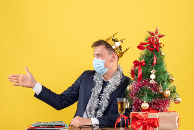 面具商务人士在圣诞树旁的桌子旁举手致意 黄色墙壁上摆放着礼物医疗面具手微笑
