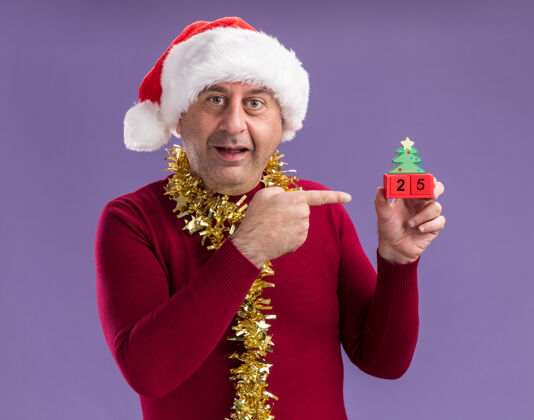 指数快乐的中年男人戴着圣诞老人帽子 脖子上戴着金属丝 手里拿着玩具方块 上面写着“25岁”的日期 用食指指着他们 站在紫色的背景上微笑着脖子五站着