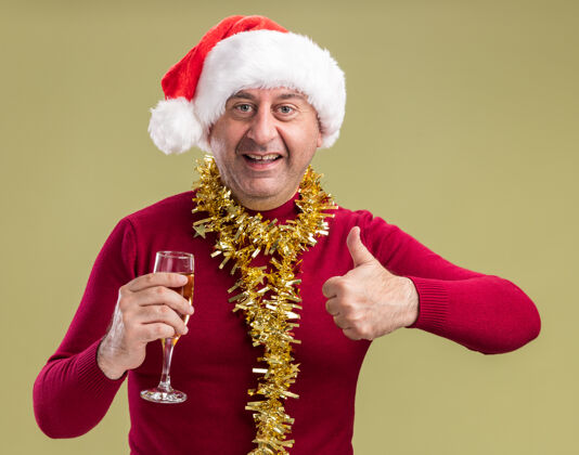 圣诞节快乐的中年男子戴着圣诞老人帽子 脖子上戴着金属丝 手里拿着一杯香槟 微笑着看着相机 竖起大拇指站在绿色的背景上香槟金属片杯子