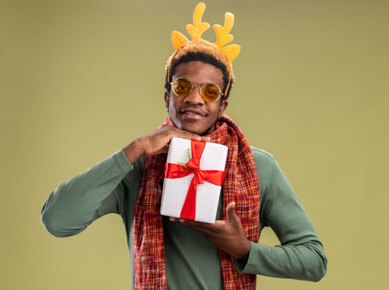 边缘一个非洲裔美国人 他脖子上戴着围巾 脖子上戴着鹿角 脖子上戴着滑稽的轮辋 手里拿着圣诞礼物 脸上带着微笑 站在绿色的背景下看着摄像机微笑抱着礼物