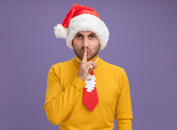 圣诞节严肃的白人年轻人戴着圣诞帽 打着领带 看着摄像机做着沉默的手势 在紫色的背景下 有着复制的空间领带沉默年轻