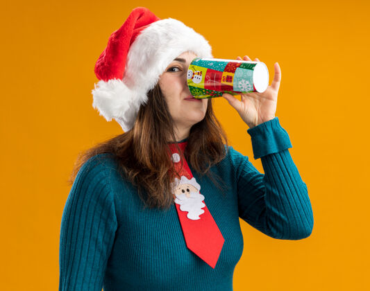 杯子高兴的成年白人妇女与圣诞帽和圣诞领带饮料从纸杯孤立的橙色背景与复制空间橙色高兴饮料