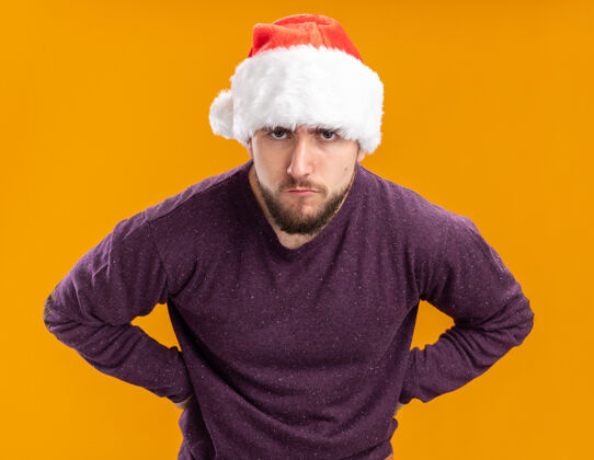 站着一个穿着紫色毛衣 戴着圣诞帽的年轻人站在橙色背景下 严肃地看着摄像机认真的年轻脸