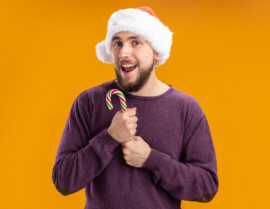 年轻人身穿紫色毛衣 戴圣诞帽的年轻人手持糖果手杖 站在橙色背景下 面带微笑地看着镜头脸抱着手杖