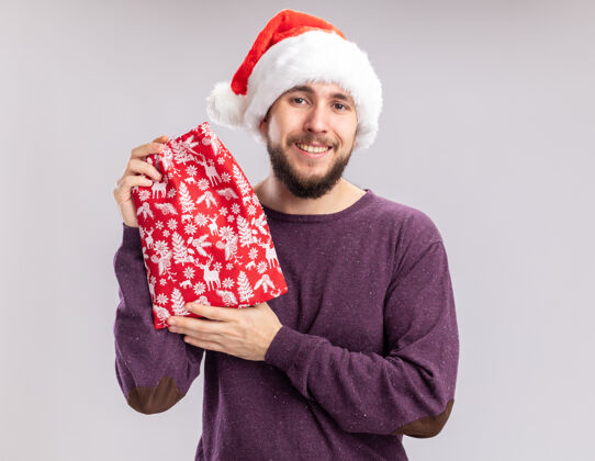 圣诞老人一个穿着紫色毛衣 戴着圣诞帽的年轻人 手里拿着红包 手里拿着礼物 面带微笑地看着镜头 站在白色的背景下脸站着毛衣