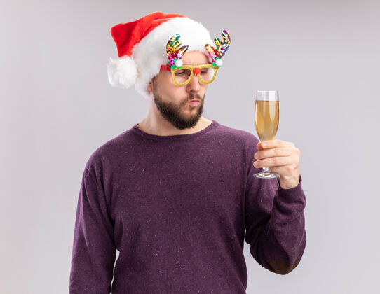 毛衣年轻人穿着紫色毛衣 戴着圣诞帽 戴着滑稽的眼镜 拿着香槟酒杯 严肃地看着它 新年假期的概念站在白色的背景上拿着眼镜帽子