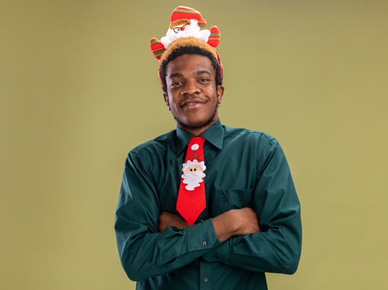 站立一个非洲裔美国人 戴着滑稽的圣诞老人戒指 打着红领带 站在绿色的背景下 开心而积极地看着镜头搞笑美国人微笑