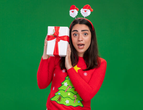 年轻焦虑的年轻白人女孩戴着圣诞老人的头带 拿着圣诞礼盒 背景是绿色的 有复印空间盒子礼物持有
