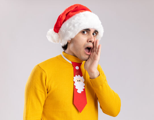嘴巴一个穿着黄色高领毛衣 戴着圣诞老人帽 打着滑稽领带的年轻人站在白色背景下 一边说着一个秘密 一边搂着嘴 一边惊讶地看着高领毛衣手臂站着