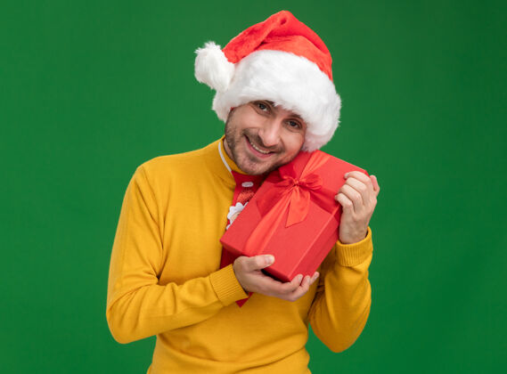 年轻快乐的年轻白人男子戴着圣诞领带 戴着帽子 拿着礼品包孤立在绿色的墙上快乐帽子领带