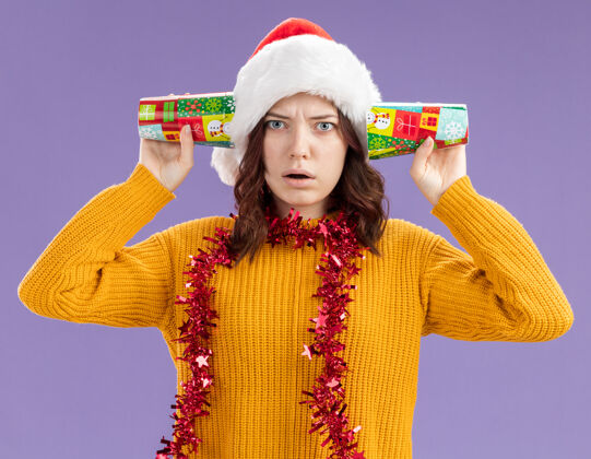 圣诞老人震惊的年轻斯拉夫女孩戴着圣诞帽 脖子上戴着花环 把纸杯放在耳朵上 紫色背景上 留有复制空间帽子年轻女孩