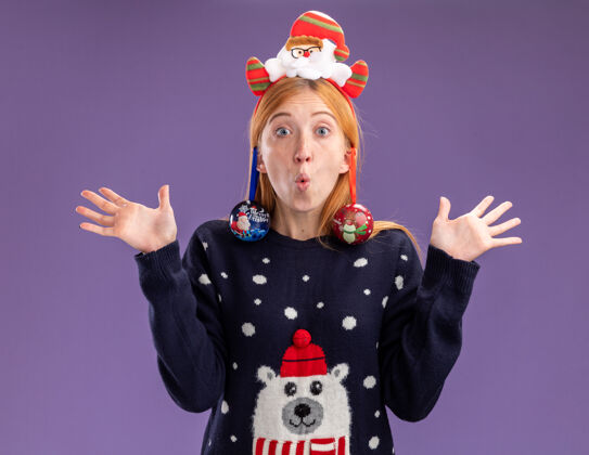 圣诞节惊讶的年轻漂亮女孩穿着圣诞毛衣 戴着圣诞发箍 耳朵上挂着圣诞球 双手分开放在紫色的背景上年轻球毛衣