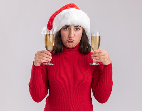 不高兴站在白色背景下 一个穿着红色毛衣 戴着圣诞帽的年轻女孩 手里拿着两杯香槟 困惑不安地看着镜头站着女孩帽子