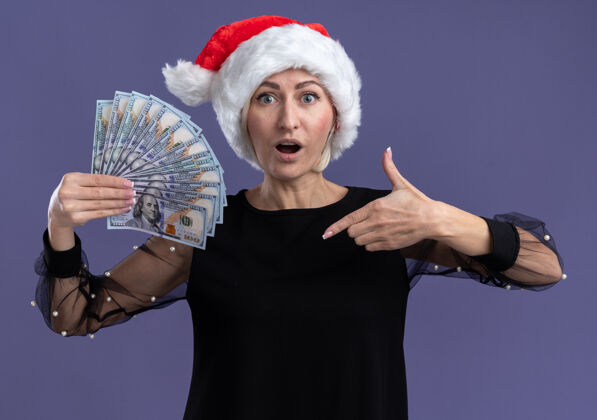 指着给人印象深刻的中年金发女人戴着圣诞帽拿着并指着隔离在紫色墙上的钱女人帽子中年