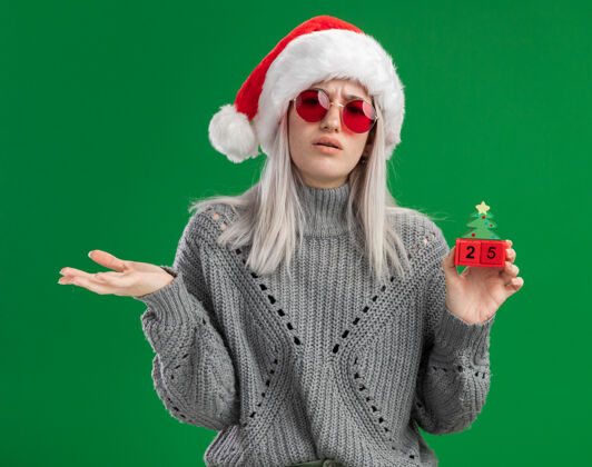 快乐年轻的金发女郎穿着冬季毛衣 戴着圣诞老人帽 戴着红眼镜 手里拿着玩具立方体 手里拿着圣诞节的日期 看起来很困惑 伸出手臂站在绿色的背景上圣诞老人圣诞快乐日期