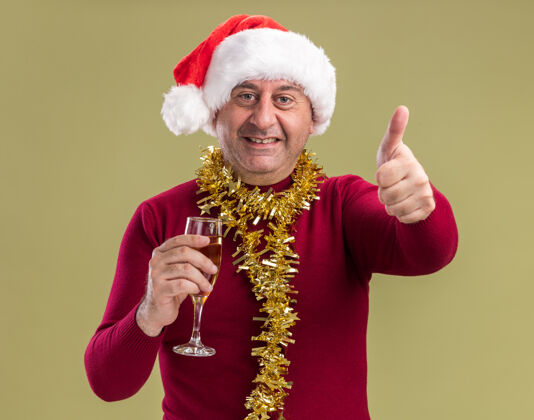 微笑快乐的中年男子戴着圣诞老人帽子 脖子上戴着金属丝 手里拿着一杯香槟 微笑着看着相机 竖起大拇指站在绿色的背景上抱着脖子圣诞老人