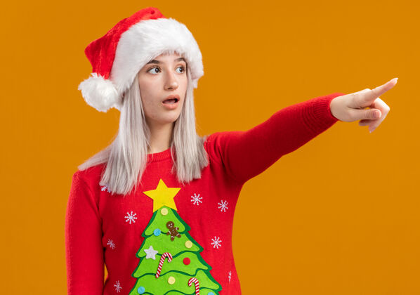 年轻人穿着圣诞毛衣 戴着圣诞帽的金发女郎惊讶地用食指指着站在橙色背景上的东西惊讶毛衣指着