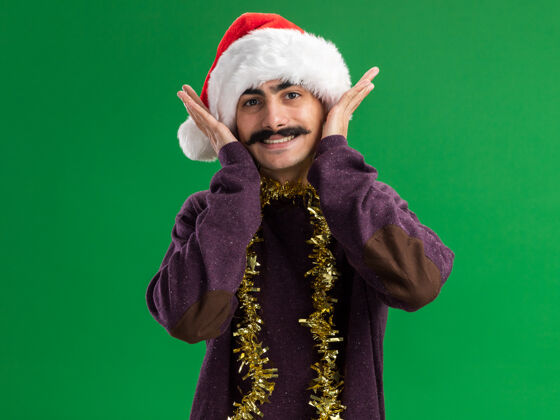 脖子年轻的留着胡子的男人戴着圣诞老人帽 脖子上戴着金属丝 站在绿色的背景下 开心地微笑着看着相机圣诞节圣诞老人快乐