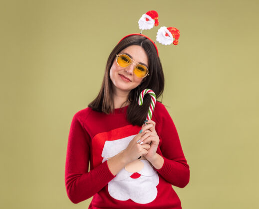 眼镜高兴的年轻漂亮的白人女孩穿着圣诞老人毛衣和头带与眼镜手持传统的圣诞糖果手杖看着相机在橄榄绿的背景与复制空间隔离头带毛衣圣诞老人