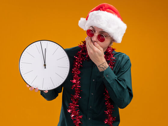 保持一个戴着圣诞帽 戴着眼镜 脖子上戴着金属丝花环的年轻金发男人 手放在嘴巴上 看着时钟 手放在橙色背景上 与世隔绝圣诞老人帽子金箔