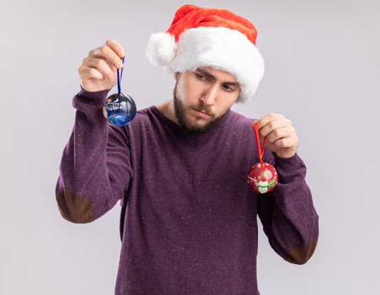 帽子一个穿着紫色毛衣 戴着圣诞帽 戴着滑稽眼镜 拿着圣诞球的年轻人站在白色背景下困惑地看着它们搞笑球眼镜
