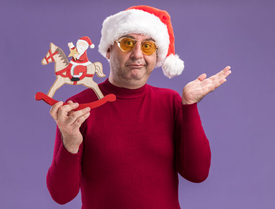 眼镜中年男子戴着圣诞老人帽 戴着黄色眼镜 展示着圣诞玩具 站在紫色的背景下困惑而不高兴地看着相机站着年龄中间