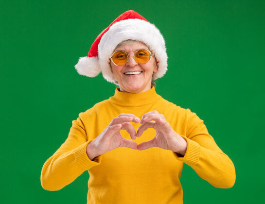 老人戴着太阳眼镜 戴着圣诞帽 面带微笑的老妇人在绿色背景上用复制空间表示孤立的心形标志心手势眼镜
