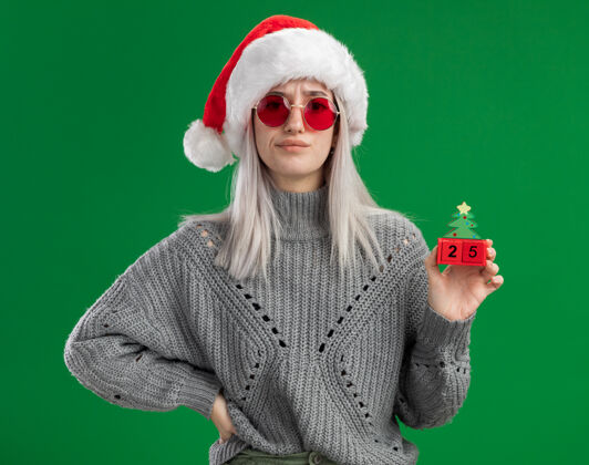 立方体年轻的金发女郎穿着冬衣 戴着圣诞帽 戴着红眼镜 手里拿着玩具立方体 手里拿着圣诞节的日期 站在绿色的背景上看着相机 很不高兴眼镜金发站着