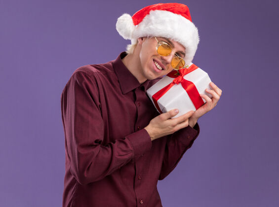 抱着微笑的年轻金发男子戴着圣诞帽 戴着眼镜 拿着礼包 用它抚摸着头 看着隔离在紫色背景上的相机感动圣诞老人包装