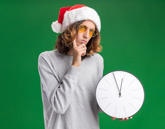 困惑戴着圣诞老人帽和黄色眼镜的年轻人举着挂钟 站在绿色的背景上困惑地看着圣诞老人年轻人钟