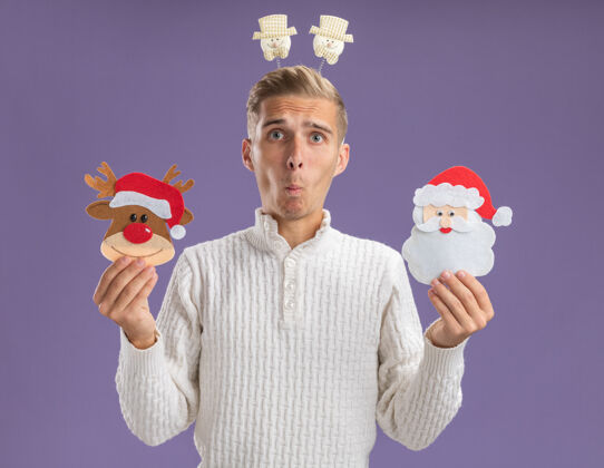 驯鹿令人印象深刻的年轻帅哥戴着雪人头带 手里拿着圣诞驯鹿和圣诞老人的纸饰品 噘着嘴唇孤立在紫色的墙上雪人印象深刻圣诞老人