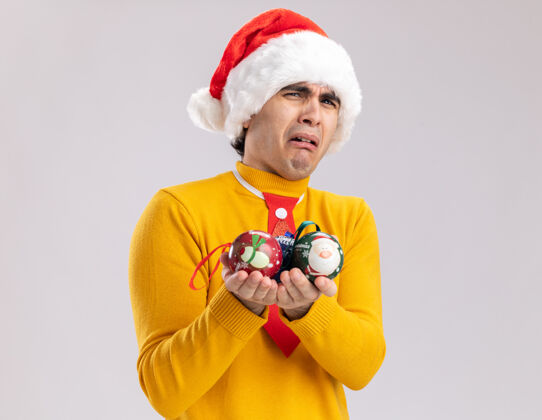 圣诞老人一个穿着黄色高领毛衣的沮丧的年轻人和拿着圣诞球的圣诞老人站在白色的背景上看着摄像机 歪歪着嘴 带着失望的表情圣诞节失望制造
