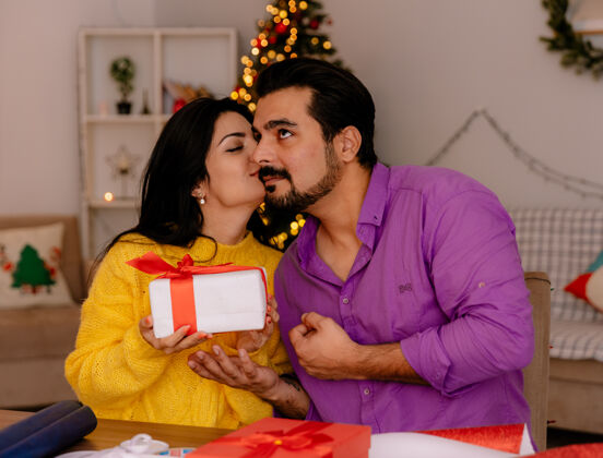 快乐一对年轻漂亮的情侣 在圣诞装饰的房间里 一个女人拿着礼物亲吻着她的男朋友 背景是圣诞树圣诞快乐圣诞树装饰