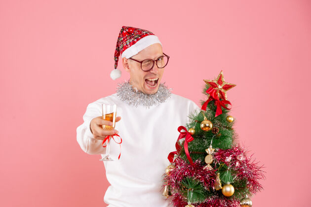 庆祝年轻人拿着小圣诞树在粉红色的墙上喝酒的正面图节日人快乐