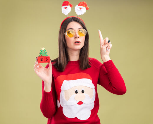 举行令人印象深刻的年轻漂亮的白人女孩穿着圣诞老人毛衣和戴眼镜的头带拿着圣诞树玩具与日期看一边指着橄榄绿的背景孤立圣诞老人毛衣年轻