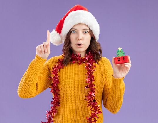 女孩惊讶的年轻斯拉夫女孩戴着圣诞帽 脖子上戴着花环 拿着圣诞树的装饰品 在紫色的背景上孤立地向上指着 还有复制空间指着年轻圣诞