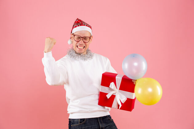 前面手持气球的年轻人的正面图 以及在粉红色墙上欢庆的礼物庆祝圣诞快乐礼物