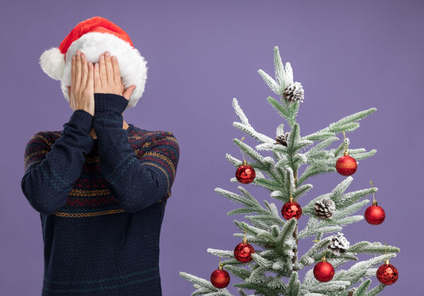 手戴着圣诞帽的白人年轻人站在装饰过的圣诞树旁 双手孤立在紫色背景上 遮住脸装饰着盖着年轻人