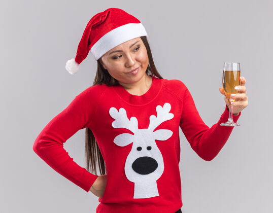 杯子想着年轻的亚洲女孩戴着圣诞帽 拿着毛衣 看着一杯香槟 把手和臀部隔离在白色背景上放着手帽子