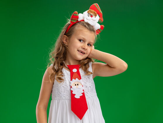 圣诞节微笑的小女孩戴着圣诞发箍 打着领带 手放在脸颊上 隔离在绿色背景上放箍脸颊