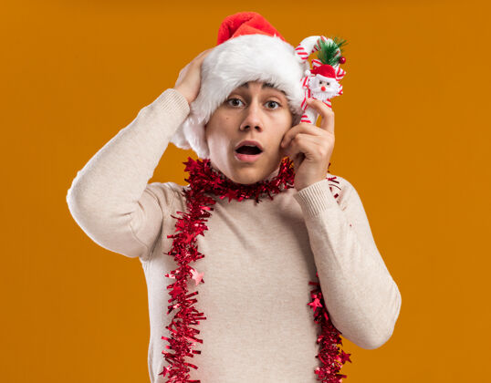 糖果关心的年轻人戴着圣诞帽 脖子上戴着花环 手里拿着圣诞糖果 手放在头上 隔离在黄色背景上关心圣诞节小伙子