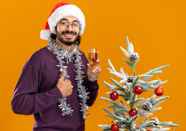 树微笑的年轻帅哥站在圣诞树旁 戴着圣诞帽 脖子上戴着花环 手里拿着一杯香槟 竖起大拇指 孤立地站在橙色背景上圣诞拇指帽子