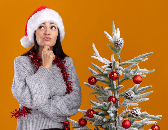 树体贴的年轻白人女孩戴着圣诞帽 脖子上戴着金箔花环 站在装饰好的圣诞树旁 抚摸着下巴 在橙色的背景上孤立地仰望脖子圣诞节年轻